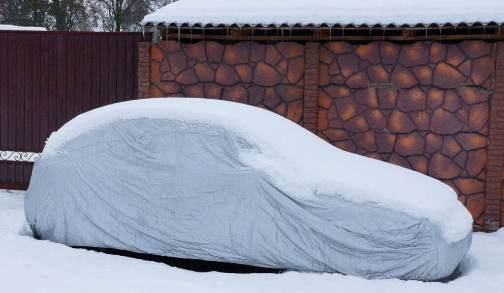 Gray tarp covering on a car in the snow by the brown fence 