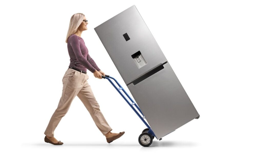 Full length profile shot of a young woman pushing a hand-truck with a new silver fridge