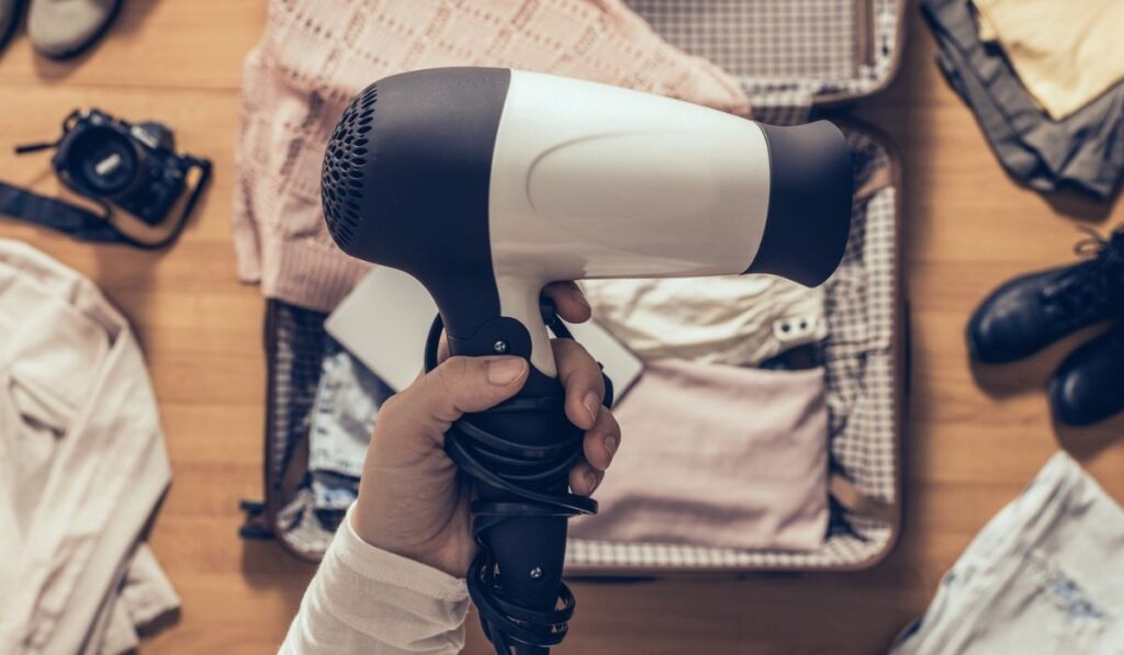 Top view on the woman holds hair dryer packing clothes in the open suitcase 