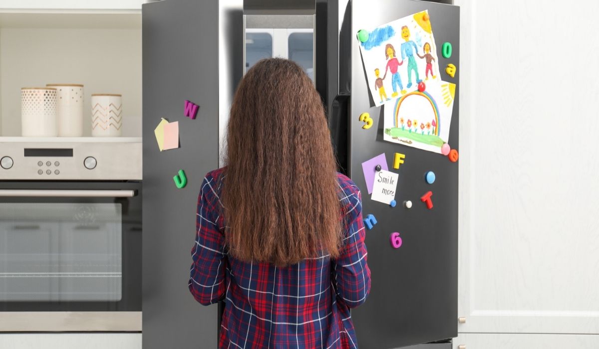 Woman opening refrigerator door with drawings, notes and magnets in kitchen